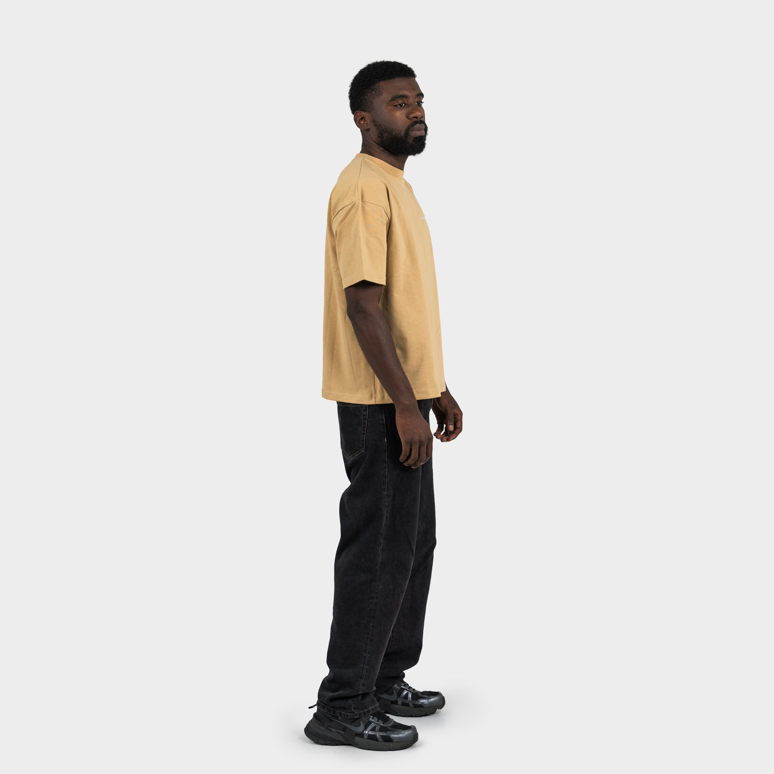 Man wearing a beige 'taze studios' t-shirt and black pants on a white background