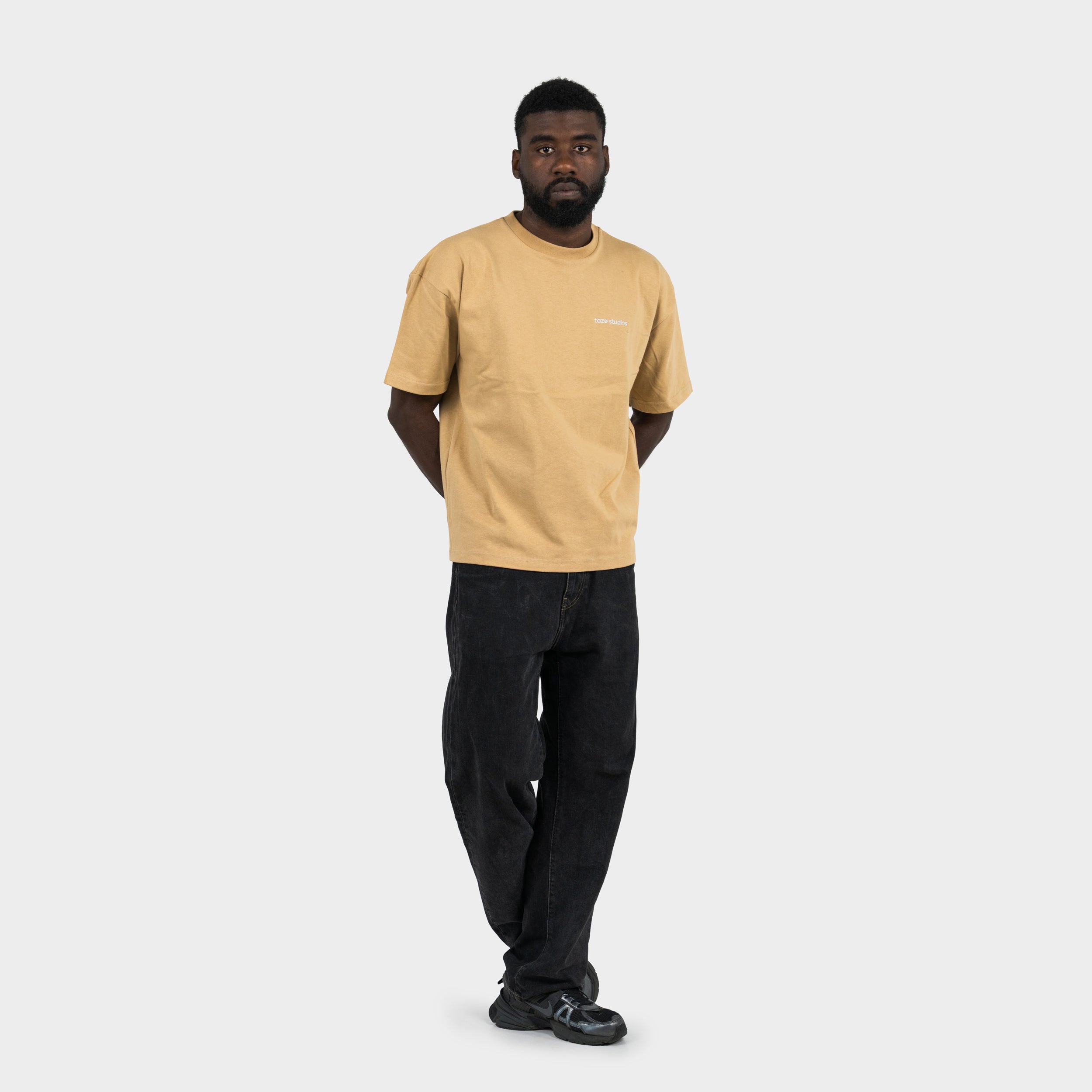 Man wearing a beige 'taze studios' t-shirt and black pants on a white background