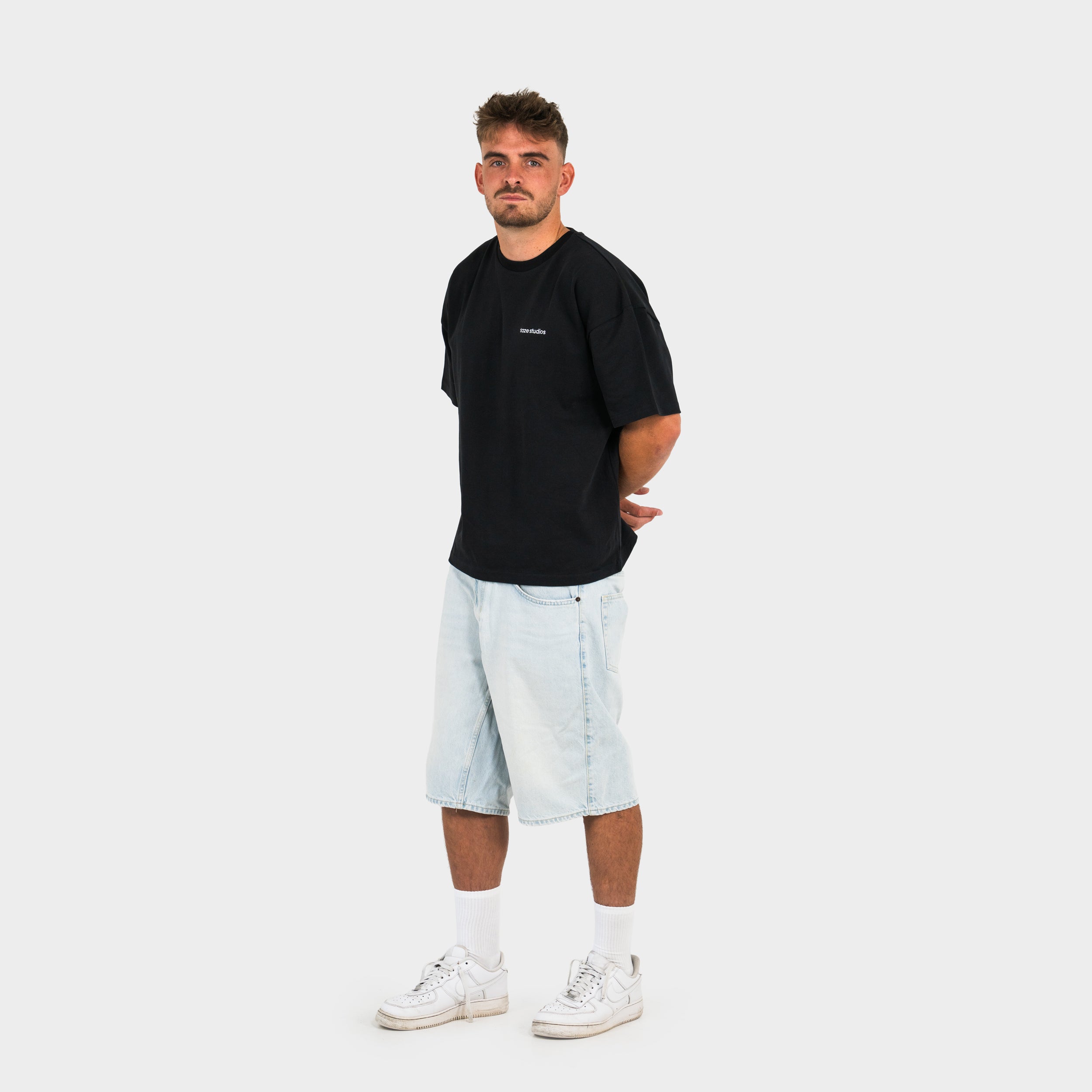 Man wearing a black 'taze studios' t-shirt and light blue shorts on a white background