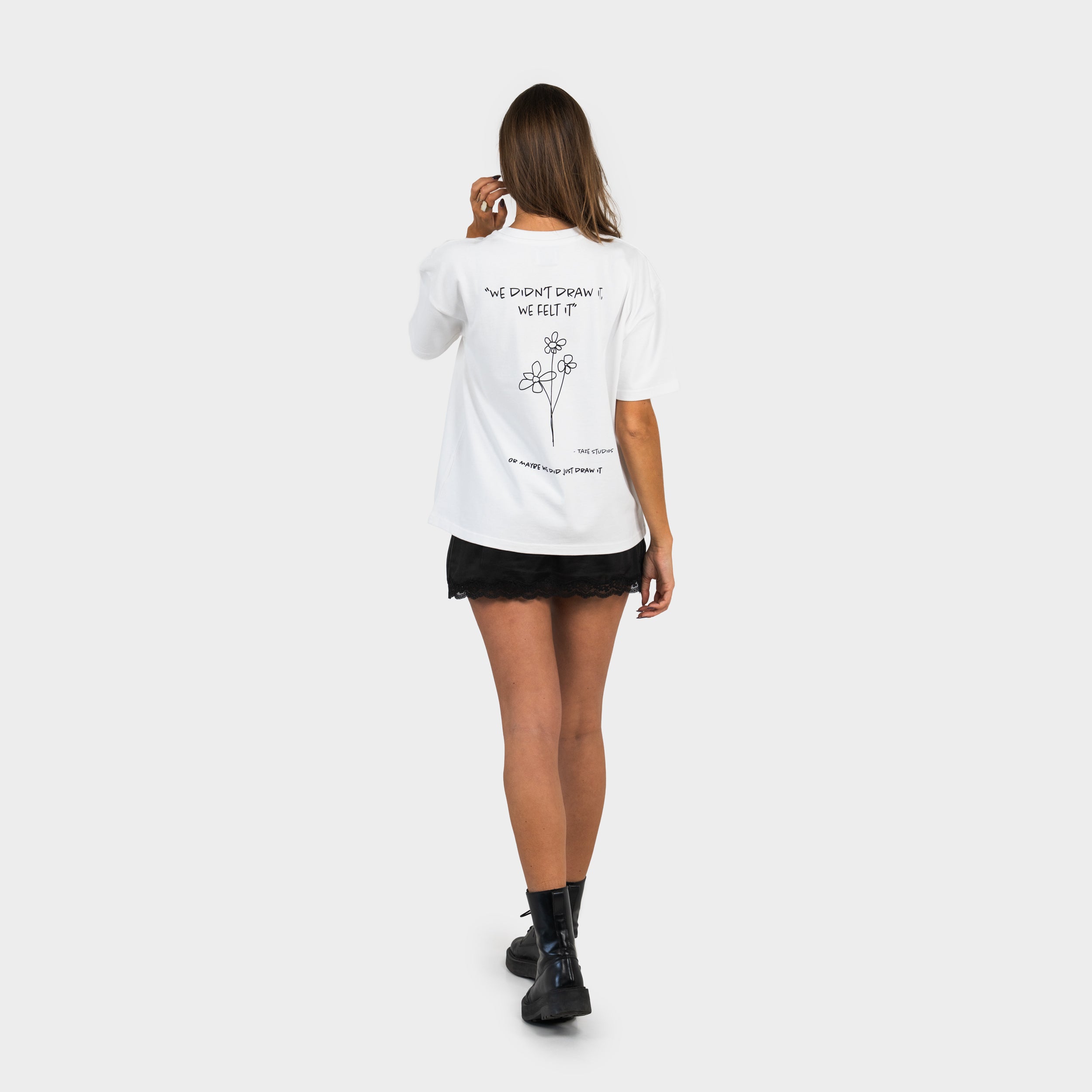 Woman wearing a white T-shirt with a 'taze studios' artwork of flowers and text on the back, standing against a white background.