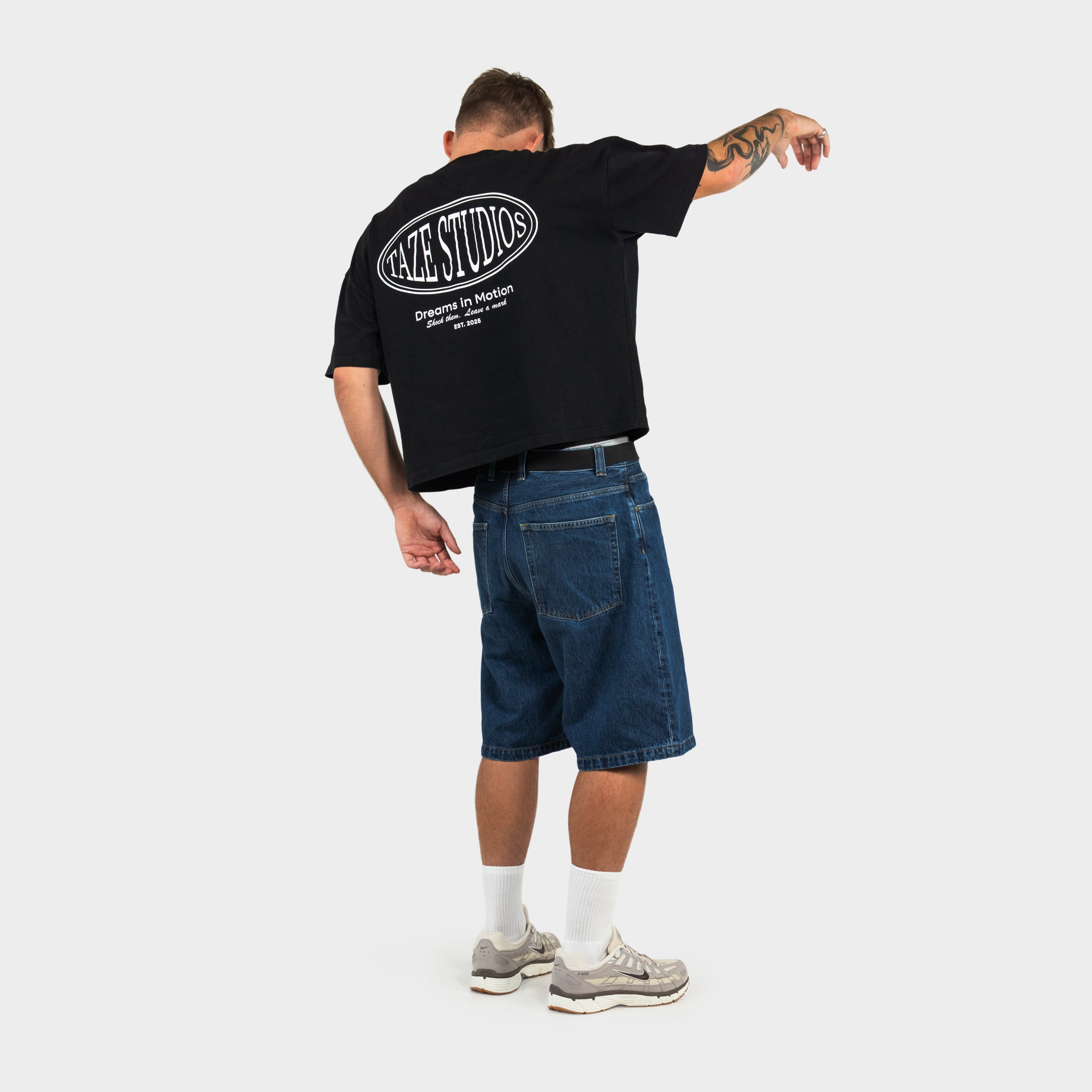 Man wearing a black boxy t-shirt with 'taze studios' artwork on the back, standing against a white background.