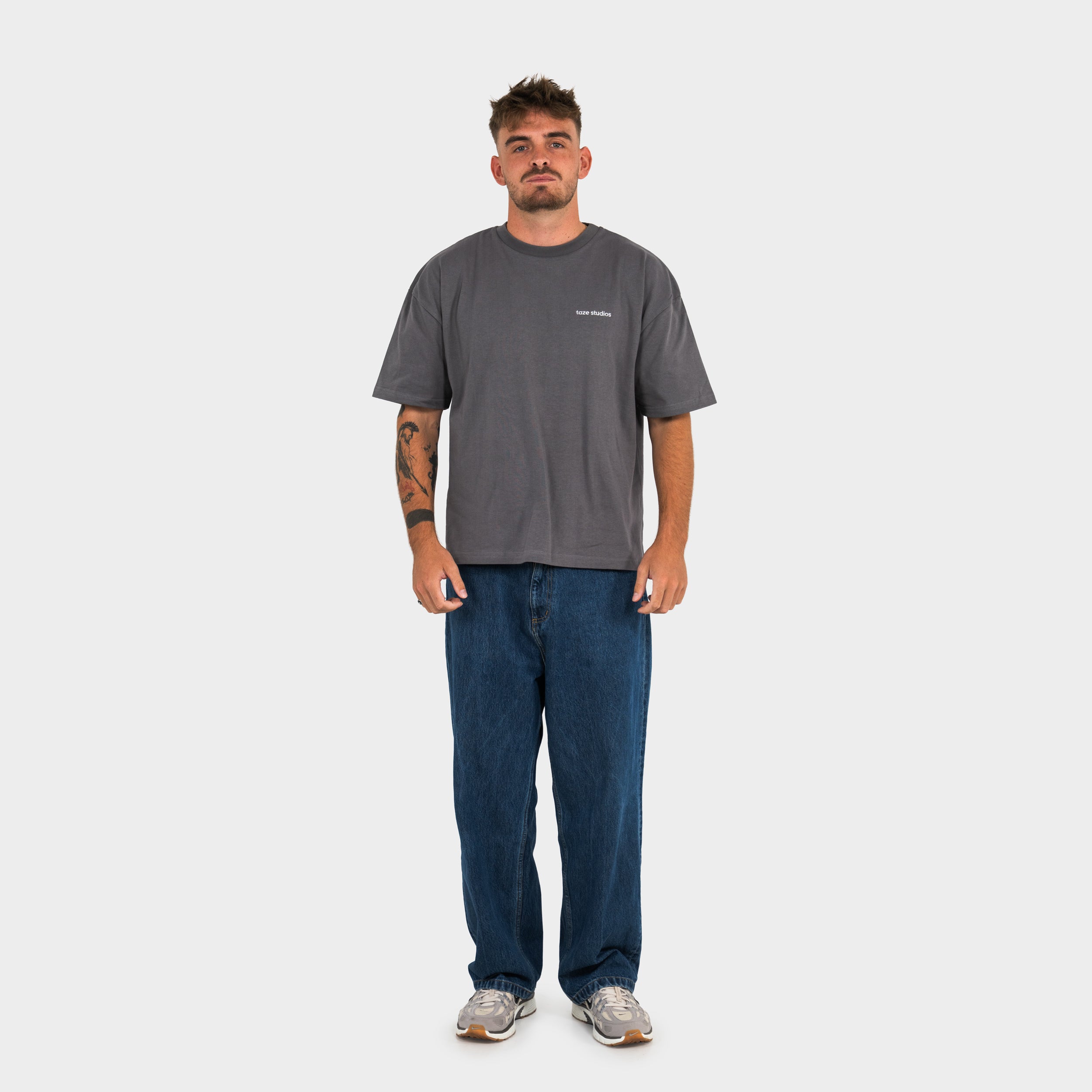 Man wearing a gray 'taze studios' t-shirt and blue jeans on a white background