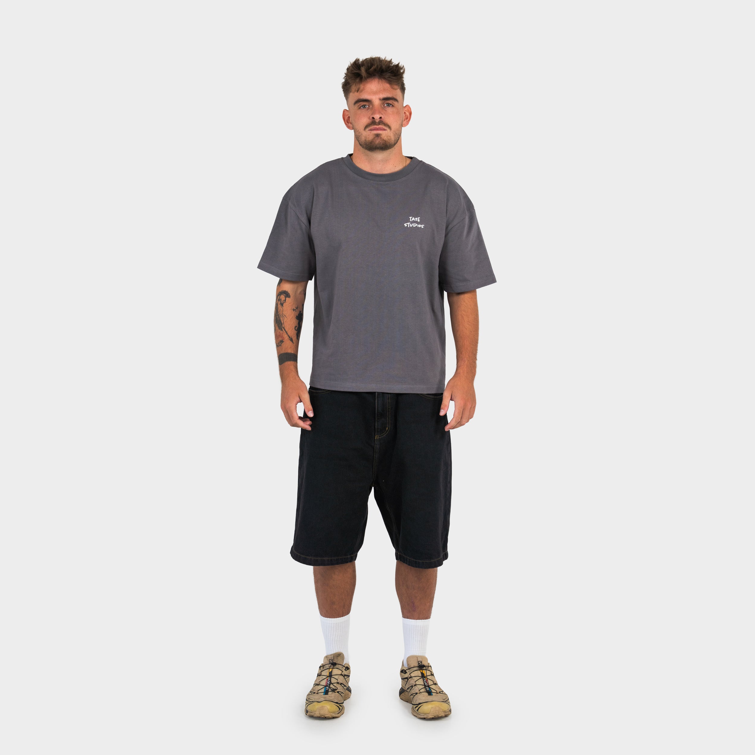 Man wearing a grey 'taze studios' T-shirt and black shorts, standing against a white background.
