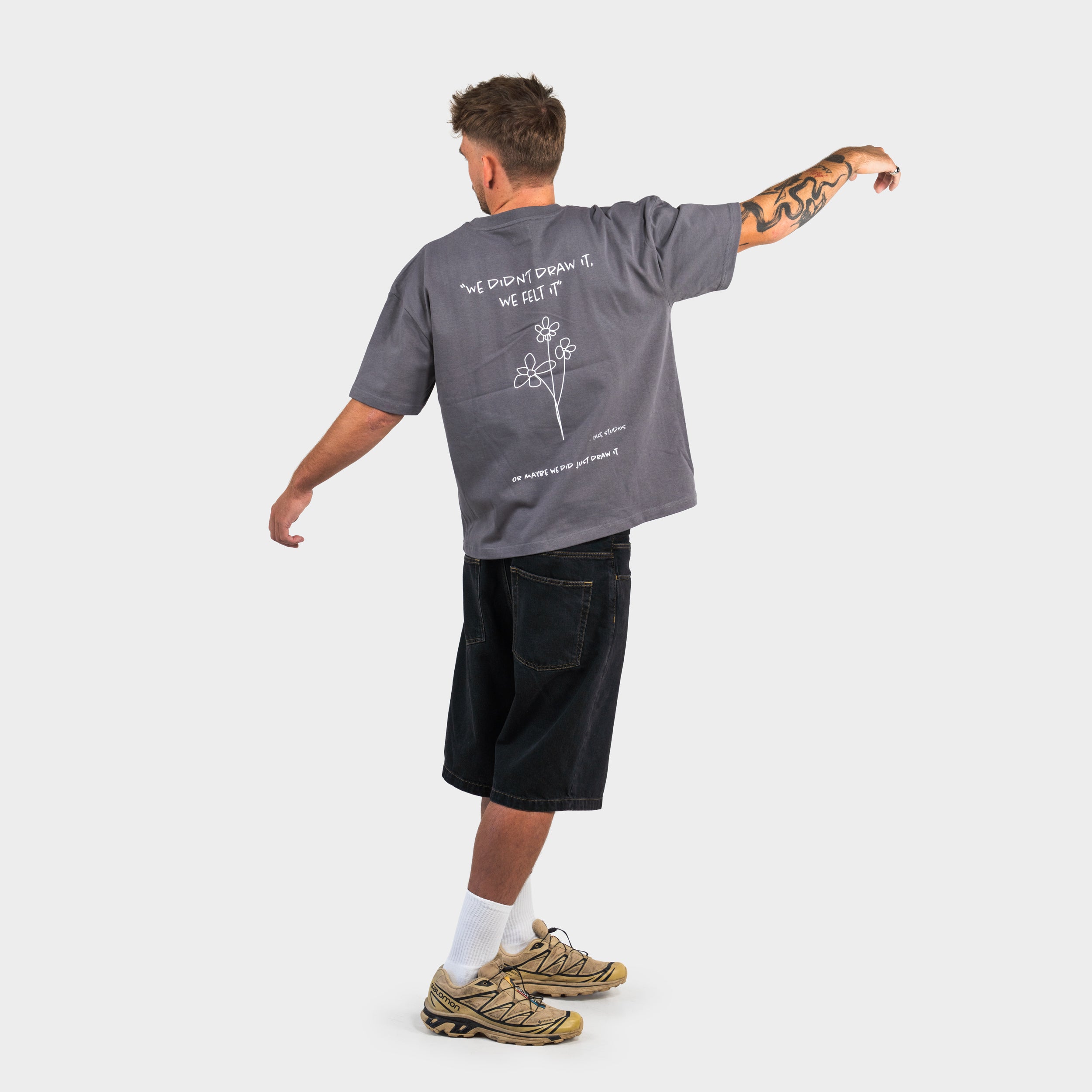 Man wearing a grey 'taze studios' T-shirt with graphics on the back and black shorts, standing against a white background.