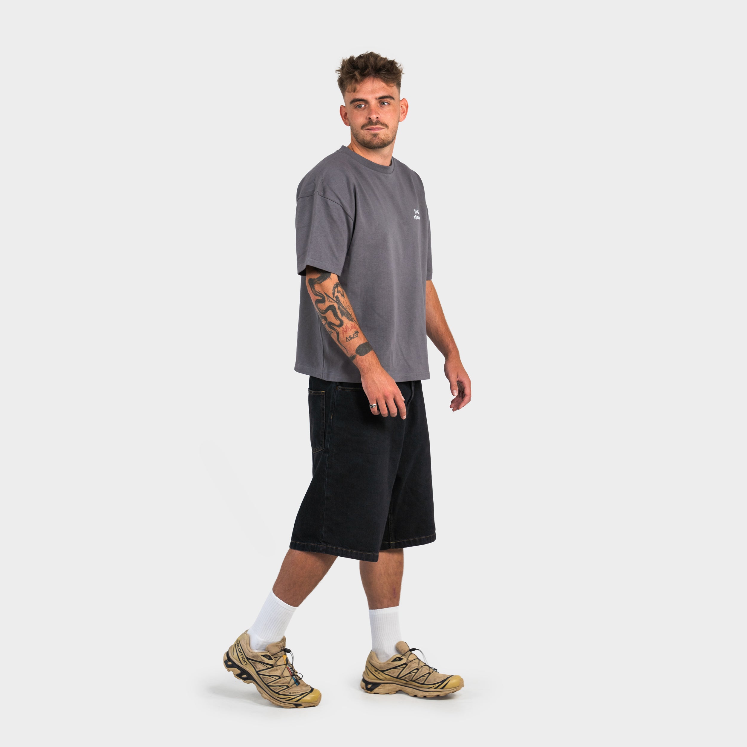 Man wearing a gray 'taze studios' t-shirt, black shorts, and beige sneakers on a white background