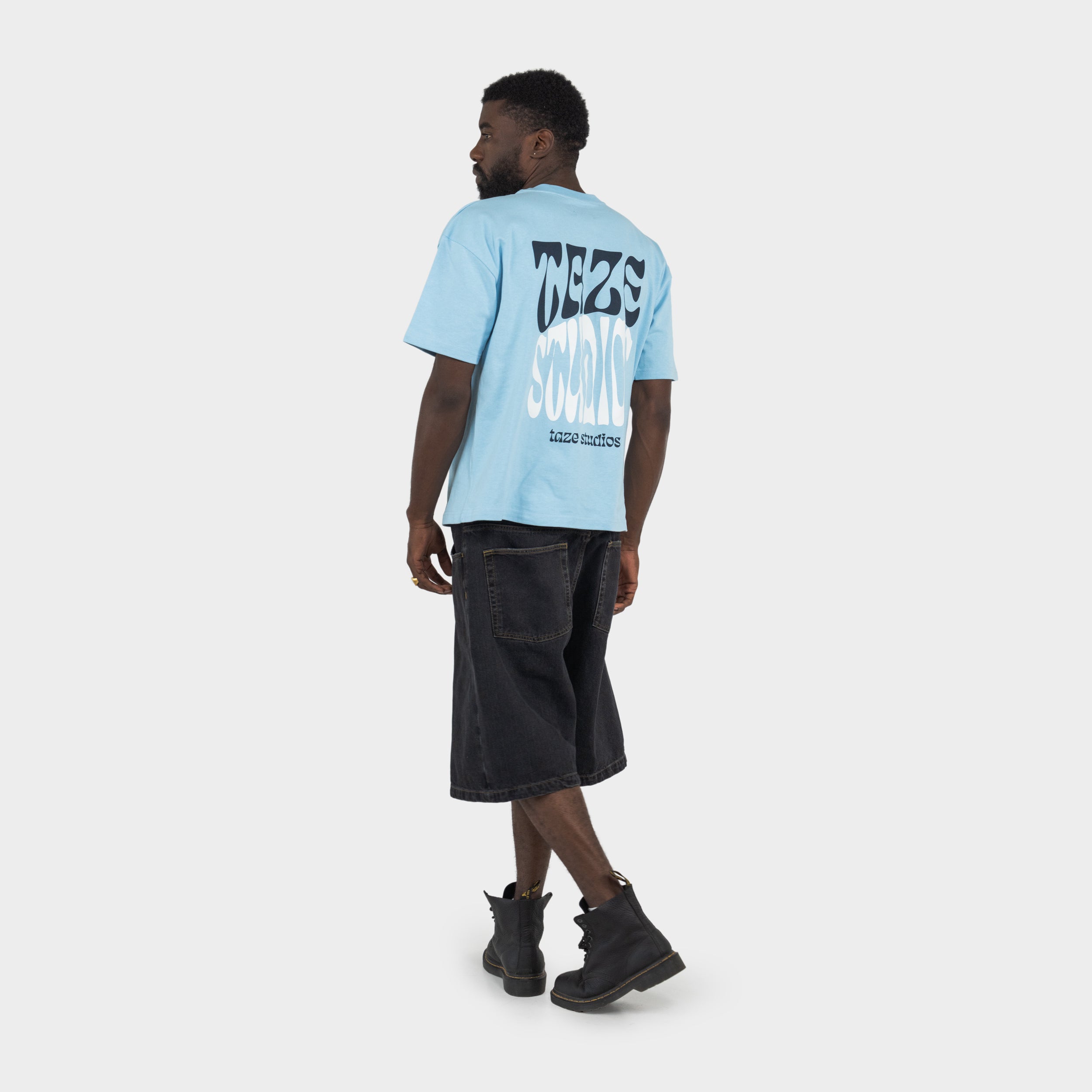 Person wearing a light blue boxy t-shirt with 'taze studios' artwork on the back, standing against a white background