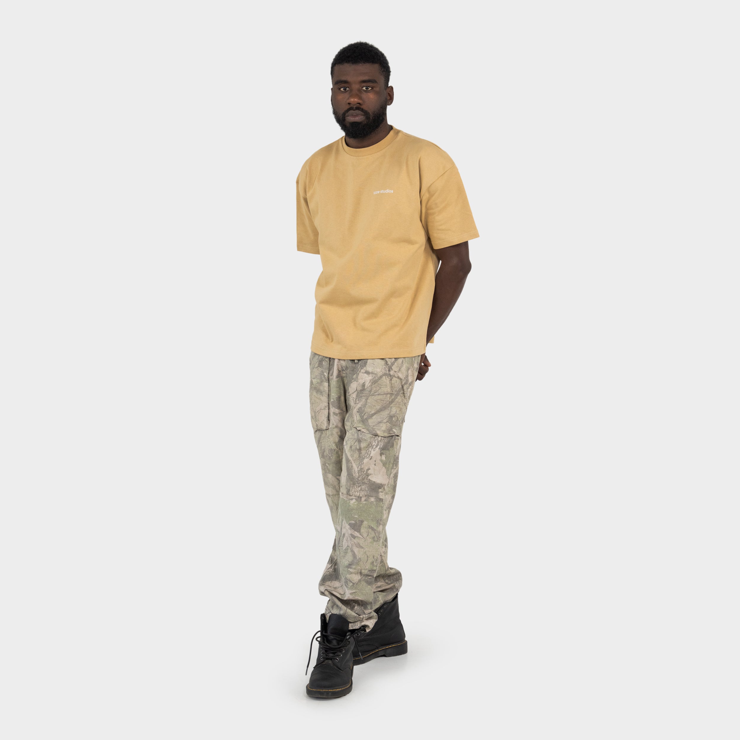 Man wearing a beige t-shirt with 'taze studios' logo on the chest and camouflage pants on a white background.