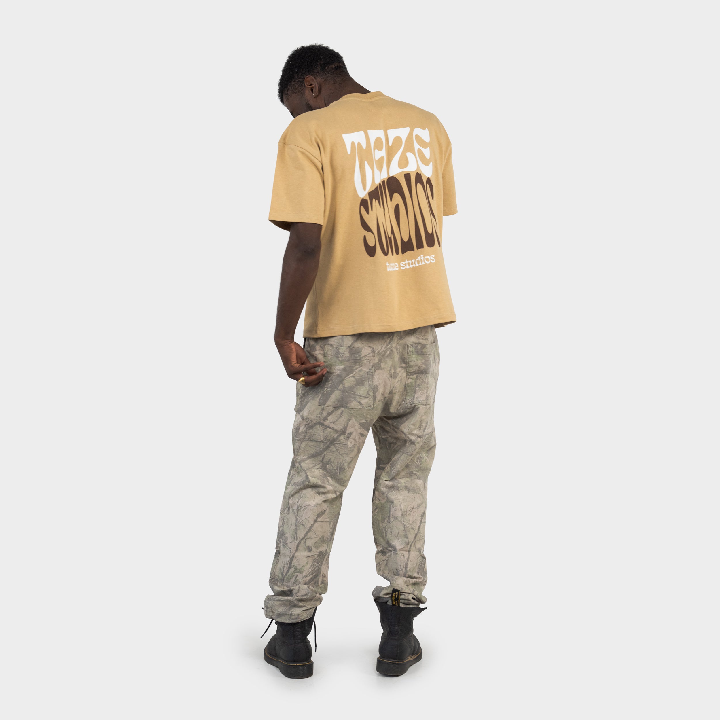 Man wearing a beige T-shirt with the 'taze studios' artwork printed on the back, and a camouflage pants, standing against a white background.