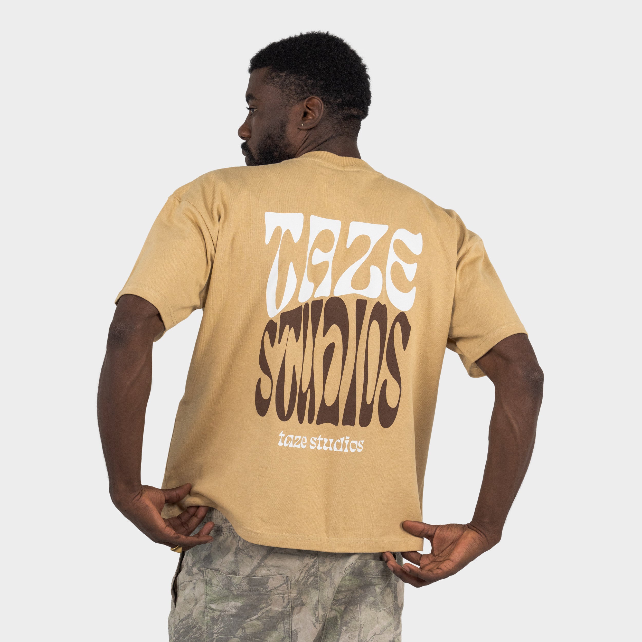 Man wearing a beige T-shirt with the 'taze studios' artwork printed on the back, standing against a white background.