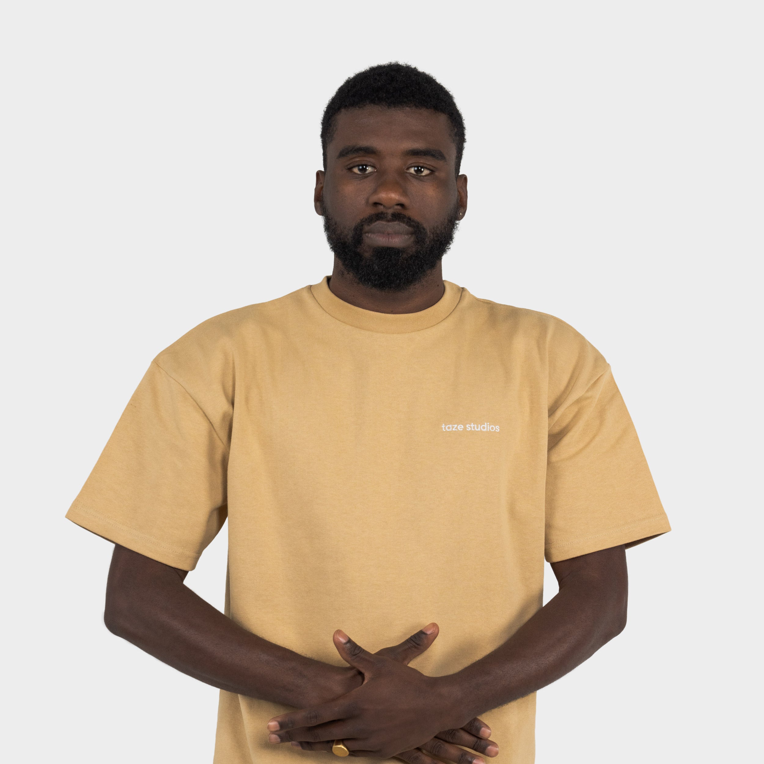 Man wearing a beige T-shirt with the 'taze studios' logo on the front, standing against a white background.