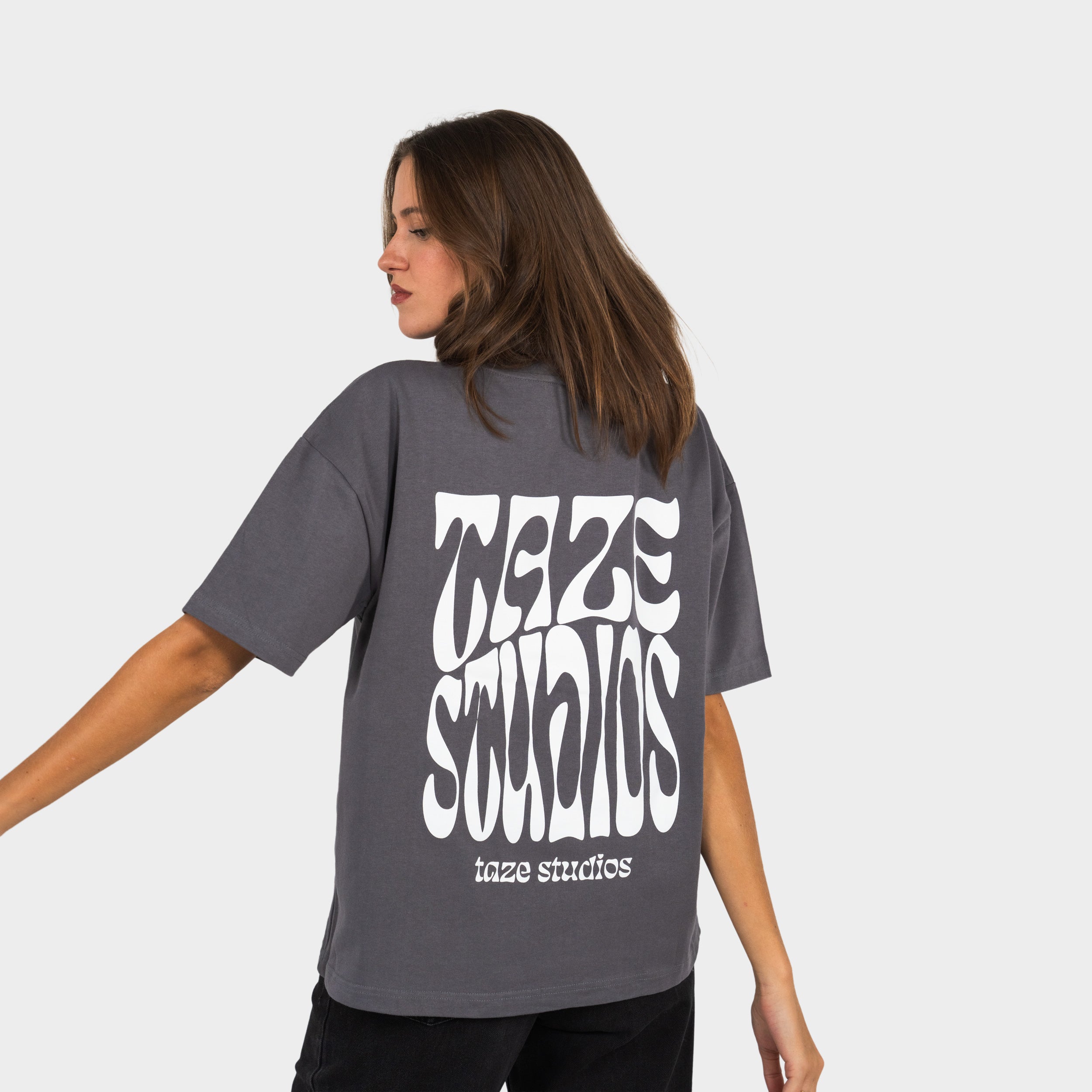 Female wearing a grey T-shirt with the 'taze studios' artwork printed on the back, standing against a white background.